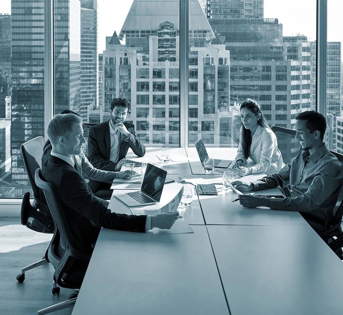 Five people in a modern office with laptops, engaged in a meeting around a long table, large windows showing a cityscape in the background.